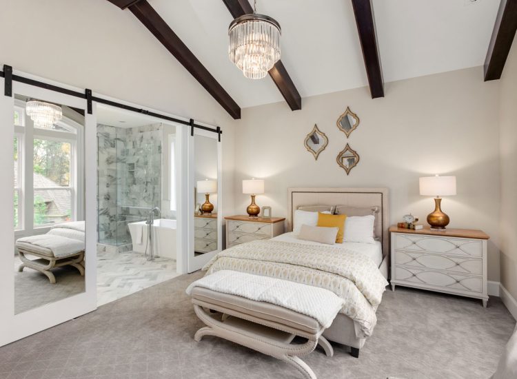 A luxurious bedroom with a vaulted ceiling, exposed dark wood beams, mirrored sliding barn doors leading to an ensuite bathroom, and elegant furnishings including a crystal chandelier.
