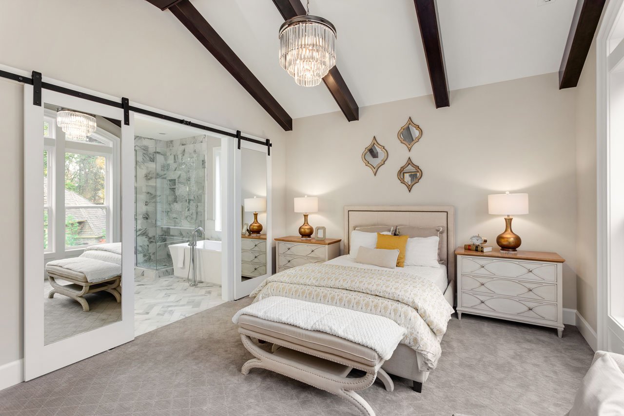 Primary bedroom suite with a vaulted ceiling and dark exposed wood beams, a crystal chandelier, and mirrored barn doors opening to an ensuite bathroom with a freestanding tub.