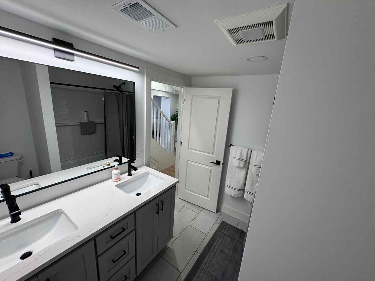 A wide shot of a fully renovated basement bathroom featuring a gray double-sink vanity, a large backlit mirror, light tile floors, and an open door leading to the hallway.