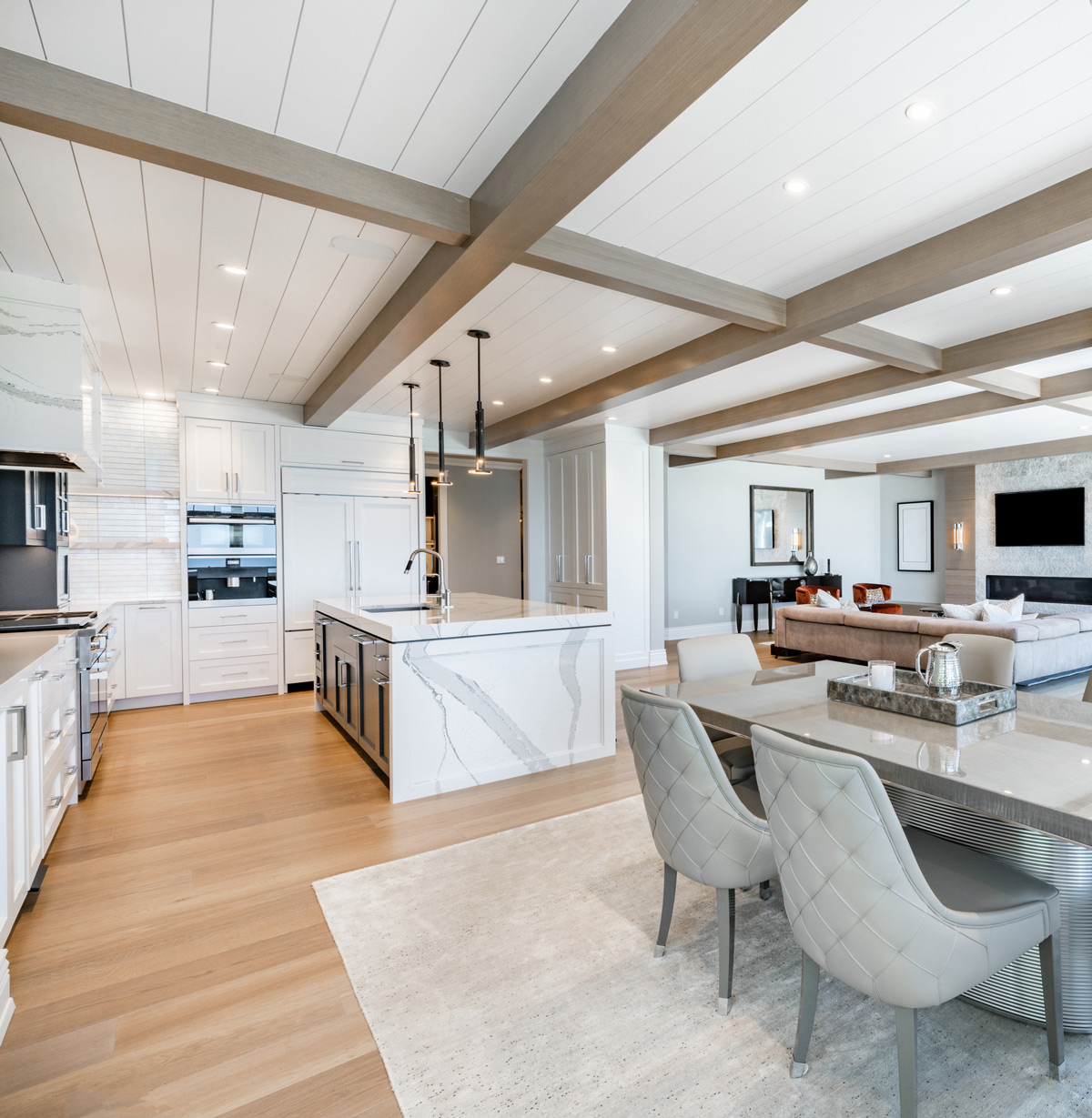 An expansive gourmet kitchen and dining space featuring a coffered wood-beam ceiling, a massive marble island, white custom cabinetry, and an open view into a comfortable living area.