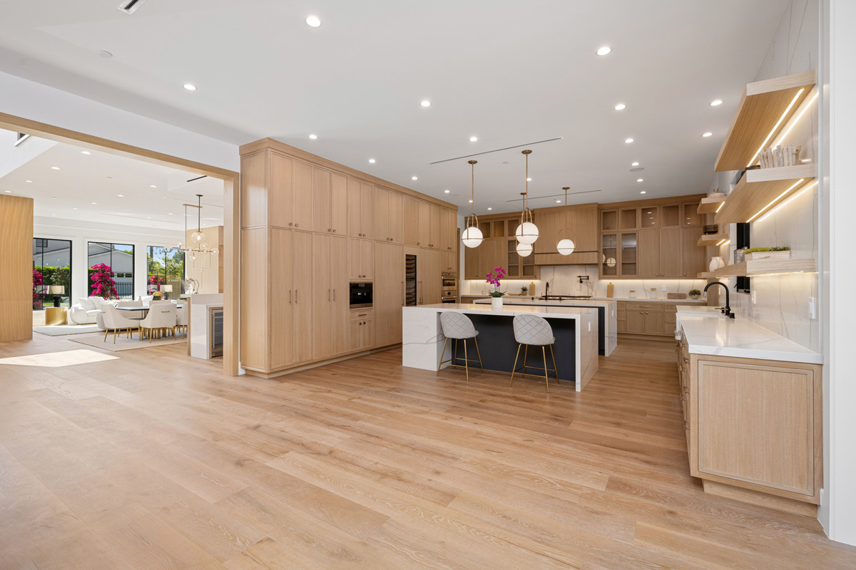 A wide-angle view of a large, modern kitchen featuring floor-to-ceiling light wood cabinets, a dual-tone island with white quartz countertops, and an open-concept layout flowing into a dining area.