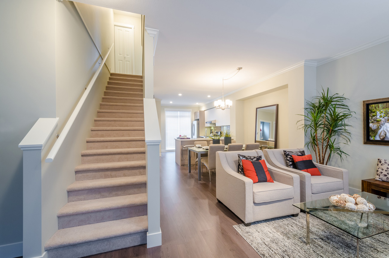 An inviting living space with hardwood floors, two neutral armchairs with colorful pillows, and a carpeted staircase leading to the upper level, all under bright recessed lighting.