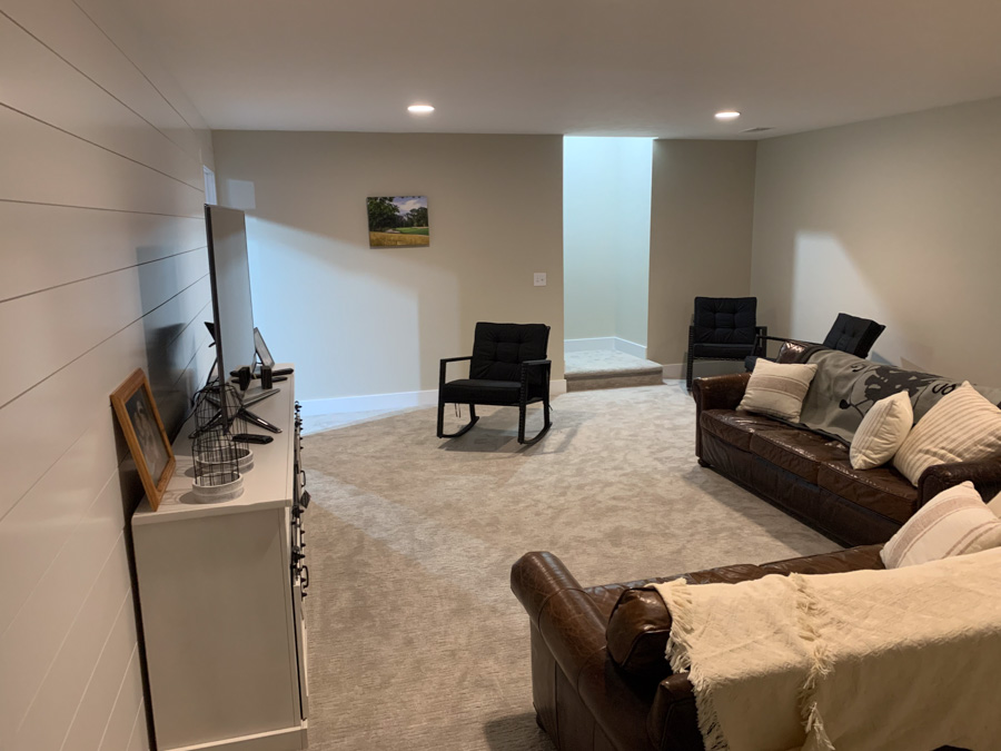 A comfortable and inviting finished basement living area with plush carpeting, leather sofas, a large TV, and a stylish white shiplap accent wall.