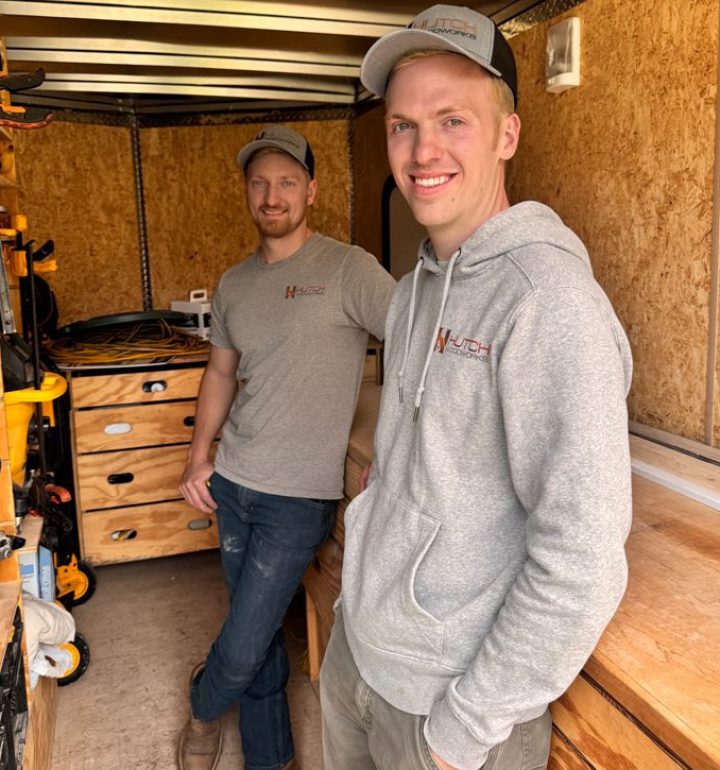 Two smiling members of the Hutch Homes construction crew standing inside a work trailer, wearing company-branded apparel and ready for a remodeling project.