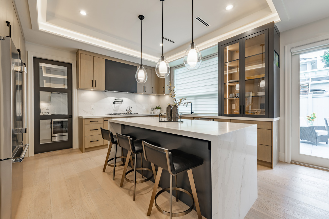 A sophisticated kitchen showcasing a mix of light wood and dark gray cabinets, a striking white marble waterfall island, modern glass pendant lights, and black leather bar stools.