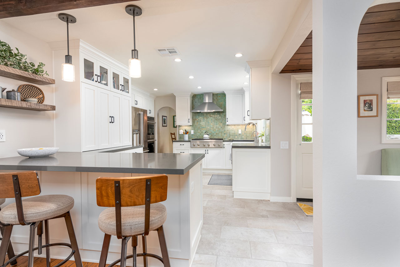 A beautiful kitchen featuring white shaker cabinets, a large island with a dark countertop and wood-back bar stools, open shelving, and a striking green tile backsplash behind the stainless steel range.