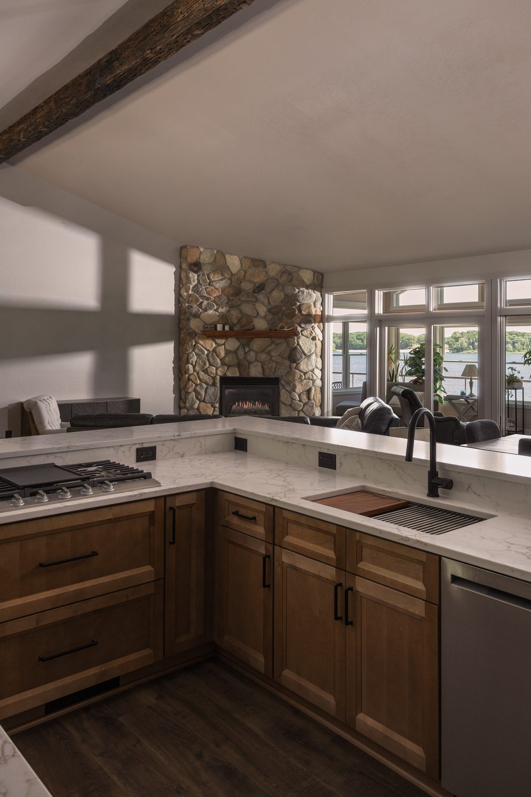 Open-concept kitchen with a river rock fireplace, exposed wood beam, and windows overlooking the lake.