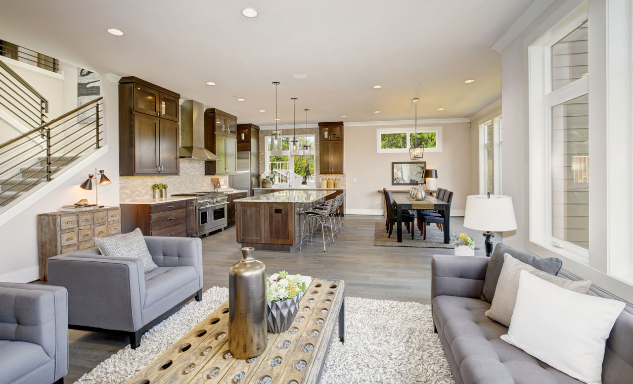 A spacious great room featuring a modern kitchen with dark wood cabinets and a large island, flowing seamlessly into a dining area and a cozy living space with gray sofas and a rustic coffee table.